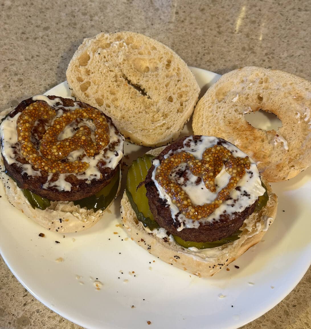 Vegan bagel burger with a plant-based patty, toasted everything bagel, vegan cream cheese, pickles, mustard, and ketchup, optionally drizzled with hot sauce.